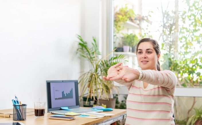 mulher sentada em cadeira de escritório alongando os braços para melhorar a postura no trabalho